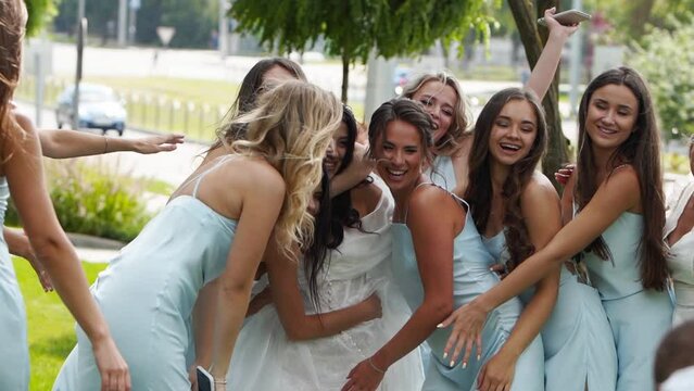 Pretty bridemaids in identical blue dresses run to hug bride together in park cheering waving hands. Woman party with friends on wedding day in elegant airy white gown and long veil. Slow motion
