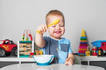 Toddler boy eats with spoon and is covered in food. Baby food, food allergies, self-feeding....
