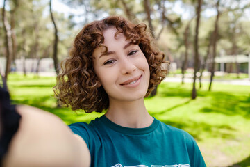 Cute redhead woman wearing green t-shirt standing on city park, outdoors taking a self portrait with smart phone. She looks at the screen and taking selfie.