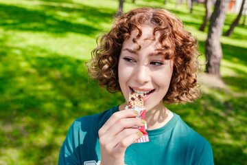 Happy redhead woman wearing tee standing on city park, outdoors biting protein or energy bar....