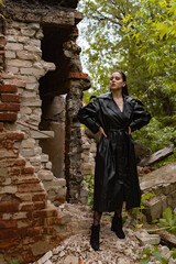 Fototapeta na wymiar a young pretty woman in a black leather coat, raincoat in a park near a destroyed building