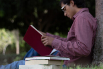 pile of books and behind them a young man reading one of those sitting on the grass in nature.