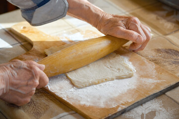 An elderly woman rolls out the dough in flour on a cutting board with a rolling pin. Woman's hands with a rolling pin.