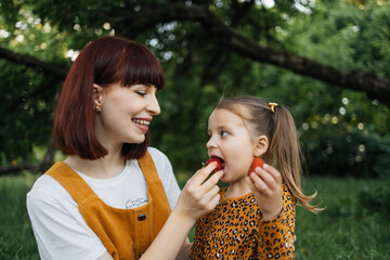 Beautiful mom and her daughter holding pieces of strawberry while resting at picnic at the park....
