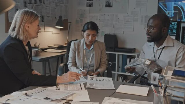 Waist-up of African American male forensics showing murder weapon in plastic bag, talking to blonde mature Caucasian female chief and Hispanic detective, sitting at desk in office at daytime