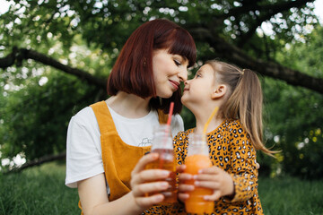 Close up view of happy caucasian mother and her cheerful daughter drinking juice in summer day outdoor in green park. Vacation and mothers day concept.