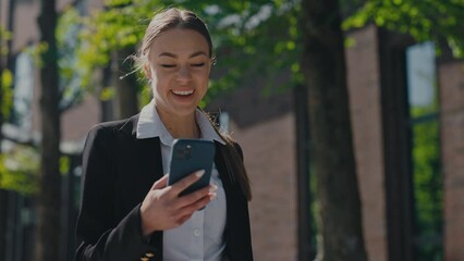 Caucasian elegant business woman receiving good news while checking emails on modern smartphone. Young lady with brown hair gesturing from happiness while walking near office center.