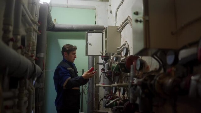 Electrician inspecting control panel with multimeter in a district heating substation. Master in uniform checking fuse box with tester probes to fix electricity. Man working in a technical room.
