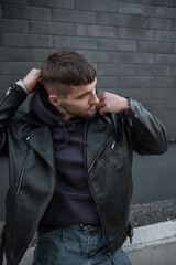 Stylish handsome model hipster guy with hair in a fashionable outfit with a leather jacket and hoodie is wearing a hood on the street near a brick black wall