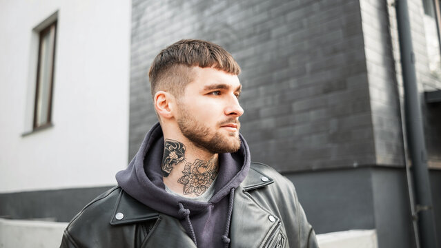 Street portrait of a handsome man hipster model with hairstyle and beard with a tattoo on his neck in a fashionable leather jacket and hoodie walks in the city