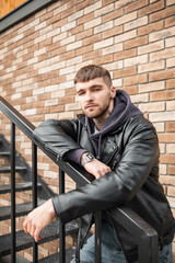 Fashionable handsome young hipster man with hair and tattoos on his neck wearing fashionable casual clothes with hoodies and a jacket stands on the stairs on the street