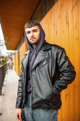 Fototapeta na wymiar Stylish hipster street guy in a fashion black leather jacket and hoodie standing next to a wooden yellow wall