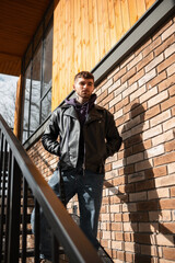 Stylish handsome young male model with hair in a fashionable leather jacket with hoodies and jeans standing on the stairs near the brick wall