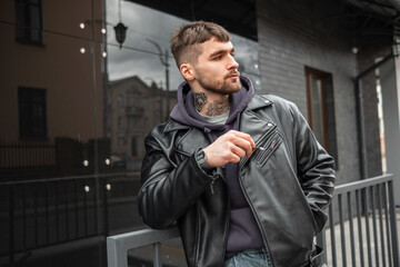 Handsome brutal hipster guy with a cigarette in fashion black outfit with leather jacket and hoodie walking in the city and smoking a cigarette - obrazy, fototapety, plakaty