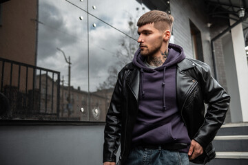 Cool handsome fashion urban man hipster with hairstyle in stylish black rock leather and hoodie walks on the street