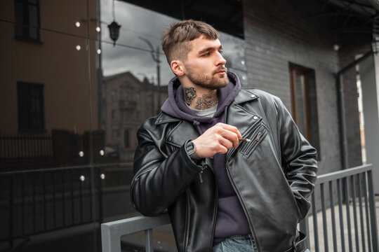 Handsome brutal hipster guy with a cigarette in fashion black outfit with leather jacket and hoodie walking in the city and smoking a cigarette