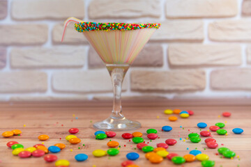 Milk cocktail in glass with drinking straw near different color round candies on wooden table. Vertical photo
