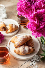 Breakfast with two plates of croissants, tea, sugar bowl and peonies flowers on brown background
