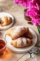 Fresh croissants, tea and pink peonies flowers for breakfast on brown background with text space