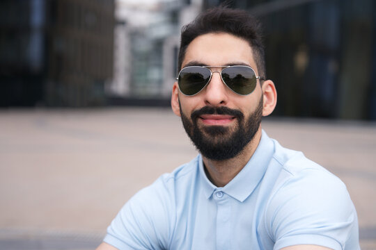 Portrait of happy positive Arab Muslim ethnic guy, young man with black beard in sunglasses and polo shirt is smiling looking at camera outdoors 