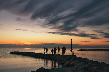 Obraz premium A small group of people watch the setting summer sun from a stone pier at Rock Harbor on Cape Cod.