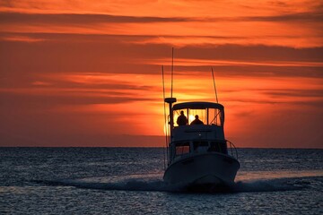 A silhouetted boat sails for harbor with the setting summer sun and a brilliant orange sky in the background.