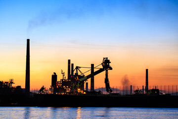 Obraz premium Dramatic sunset behind smokestacks and industrial crane in factory harbor