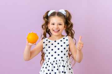 Obraz na płótnie Canvas A little girl holds an orange and gives a thumbs up. A child in a polka dot dress holds a fruit on a pink isolated background.