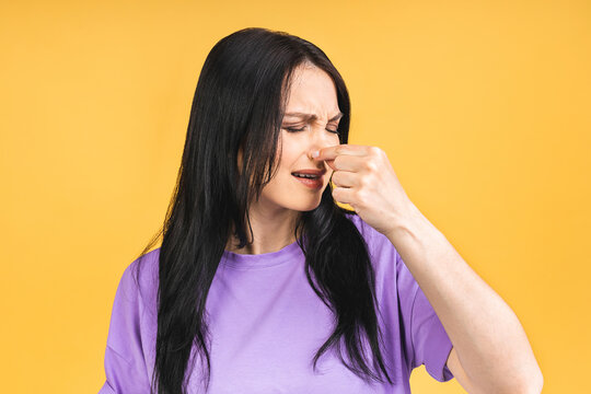 Portrait Of Frustrated Woman Pinch Nose With Disgust On Face Due To Bad Smell Isolated Over Yellow Background.