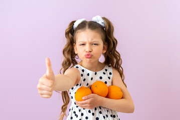 The child holds oranges, smiles broadly and gives a thumbs up. Fruits and vitamins for children. A beautiful girl in a polka dot dress on a pink isolated background.