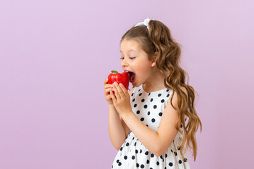 A little girl is about to bite off a piece of pepper on a pink isolated background. A beautiful...