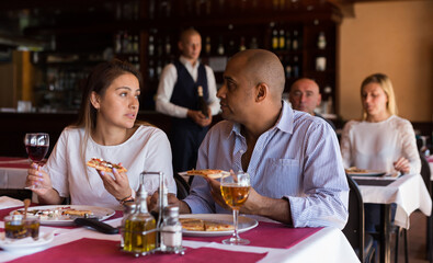 Young hispanic couple enjoying dinner with delicious pizza and wine in cozy italian restaurant