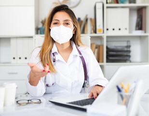 Woman doctor in medical mask is working at laptop in clinic