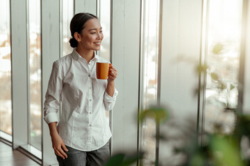 Fototapeta premium Portrait of asian business woman drinking coffee standing near windows at office. Blurred background