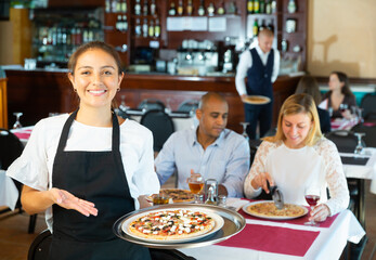 Portrait of smiling attractive hispanic waitress with serving tray warmly welcoming in cozy pizza restaurant