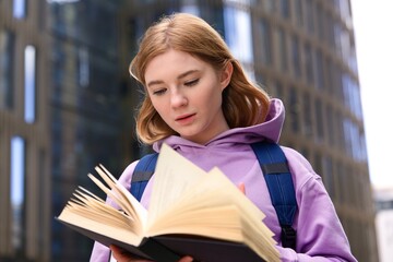 Young beautiful blonde woman, college or university academic student girl or school pupil is studying, preparing to lesson, reading book or textbook outdoors at campus with backpack 
