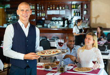 Portrait of smiling elegant waiter holding plate with appetizing pizza, greeting guests in cozy pizzeria