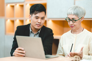 Fototapeta na wymiar Young Asian business man in formal clothing showing something on digital tablet to senior female boss and manager wearing eyeglasses while in a meeting and training on technology
