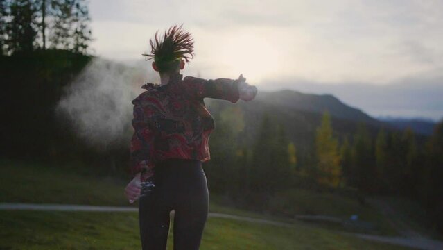 Slow motion close up of a young woman dancing on a wooden platform at sun set, throwing white powder. Alpine mountains and an orange cloudy evening scenery in the background. Gimbal Shot in 4K.