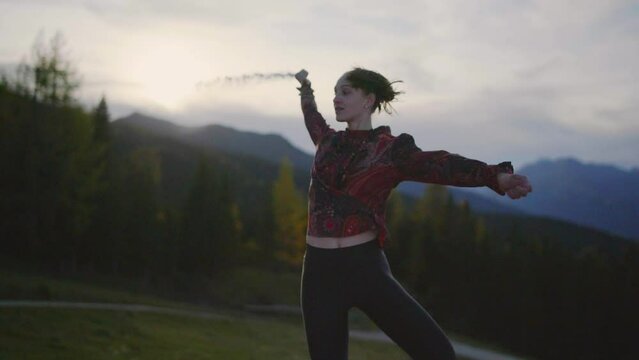 Slow motion close up of a young woman dancing on a wooden platform at dusk, throwing white powder. Alpine mountains and an orange cloudy evening scenery in the background. Gimbal Shot in 4K.