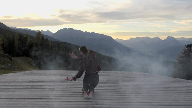 Young woman sitting and dancing on a wooden platform in the mountains at dusk. Alpine mountains and an orange cloudy evening sky scenery in the background fog coming into the frame. Gimbal Shot in 4K.