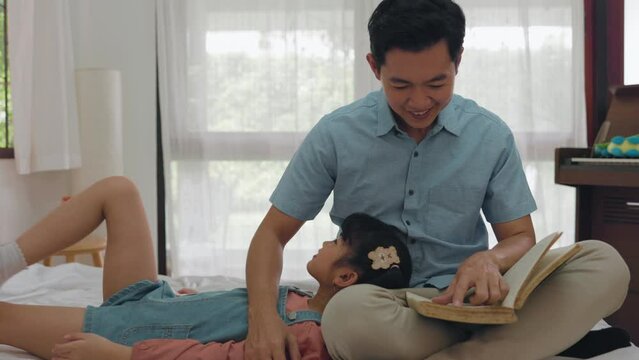 Asian adult dad reads fairy tales to cute little daughter in bed at home
