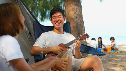 Asian couple enjoying together camping on ther beach male playing ukulele having fun and freedom on summer vacation people lifestyle activity on weekend concept.