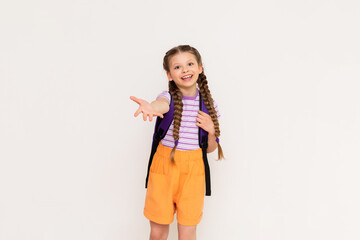 A little girl with a school backpack on her back stretches her hand forward. A beautiful child with pigtails and shorts on a white isolated background.