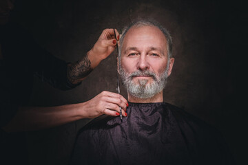 Portrait of woman hairdresser cutting hairs of elderly man looking at camera against dark background.