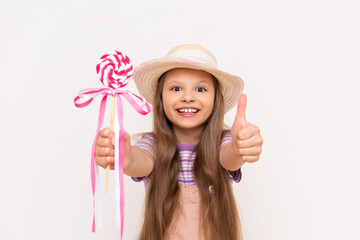 A little girl shows a lollipop and smiles. A child in a sundress and a summer hat on a white isolated background shows a thumbs up.