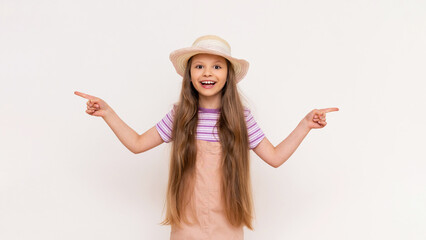 A beautiful girl points sideways at your advertisement. A little girl in a summer sundress and a straw hat on a white isolated background.