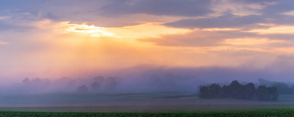 The first rays of the sun break through the fog and clouds and illuminate the green field