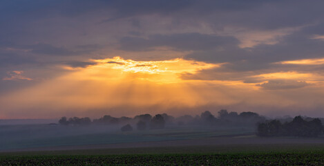 The first rays of the sun break through the fog and clouds and illuminate the green field