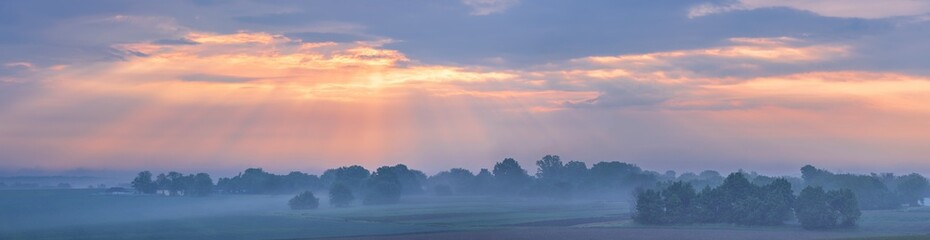 The first rays of the sun break through the fog and clouds and illuminate the green field